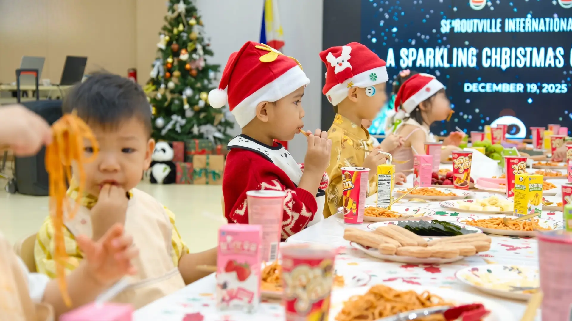 Preschool children wearing cute Santa hats eating spaghetti and snacks at a long decorated table during the annual holiday celebration event at Sproutville International School.