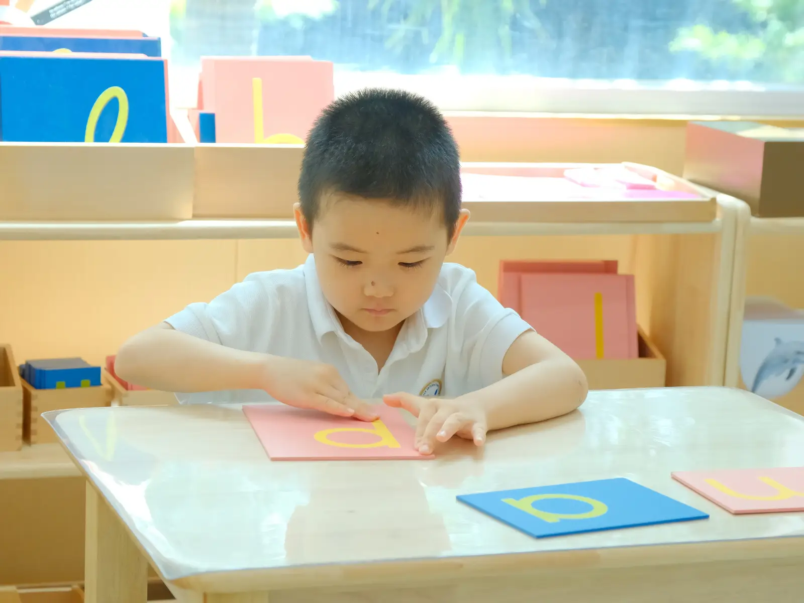 Young student at Sproutville International School practicing Montessori language skills by tracing tactile sandpaper letters at a wooden desk.