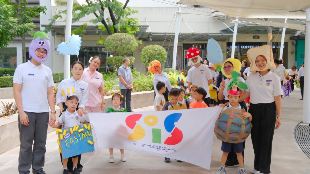 Teachers and students wearing creative food costumes holding a school banner during the outdoor Nutrition Month parade at Sproutville International School.