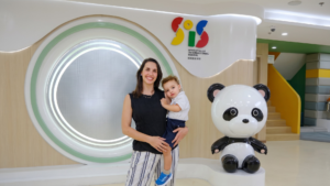 A happy parent holding her young toddler student in the welcoming lobby of Sproutville International School next to the panda mascot.