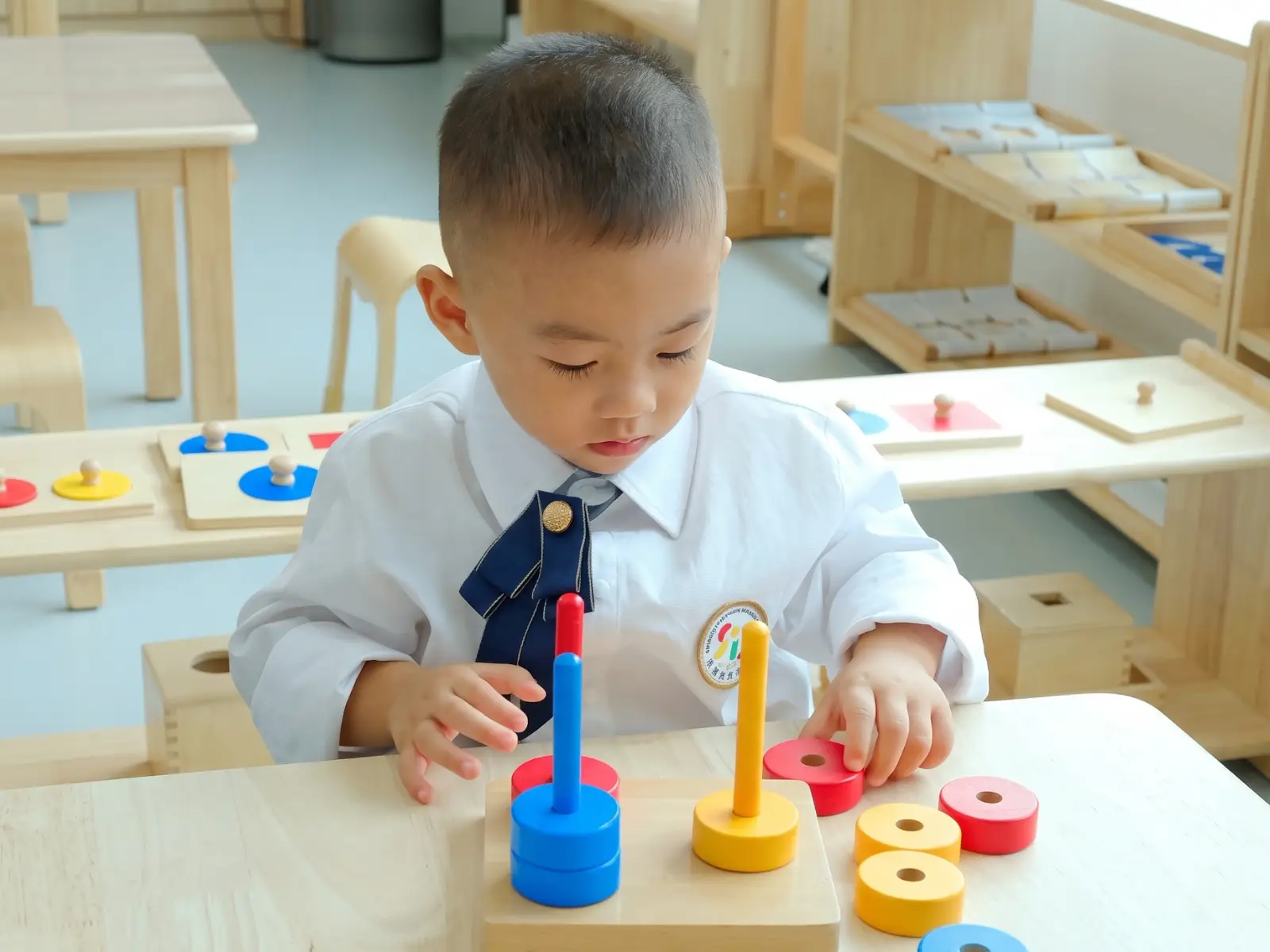 A focused young boy in uniform at Sproutville International School developing fine motor skills using colorful wooden stacking rings in a Montessori classroom.