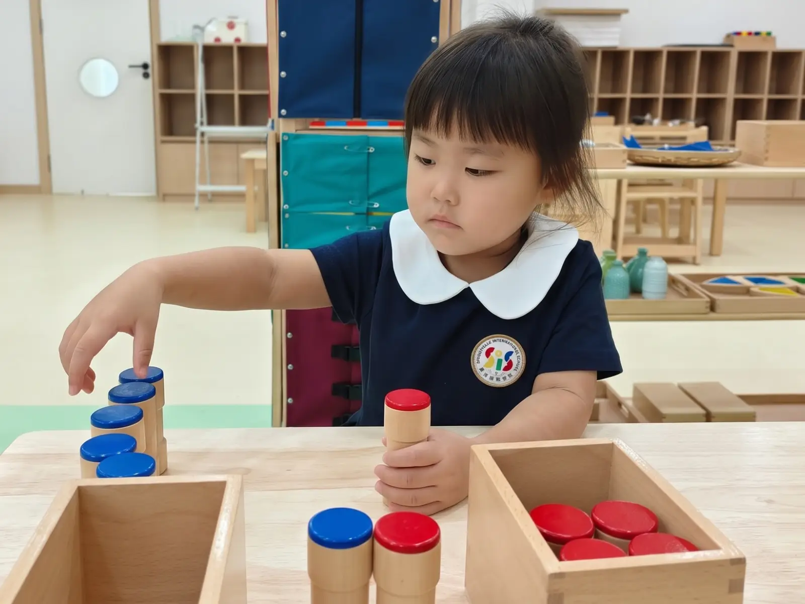 A focused preschool girl at Sproutville International School engaging in a Montessori sensory lesson by carefully arranging red and blue wooden knobless cylinders.